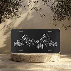 May include: Black metal license plate with a white outline of a mountain range and trees.