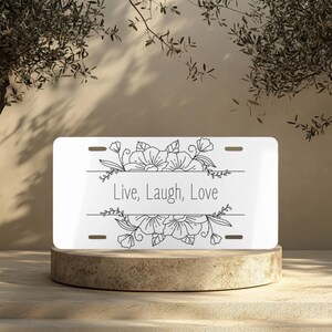 Puede incluir: Placa de matrícula personalizada blanca con un diseño floral negro y el texto "Live, Laugh, Love".