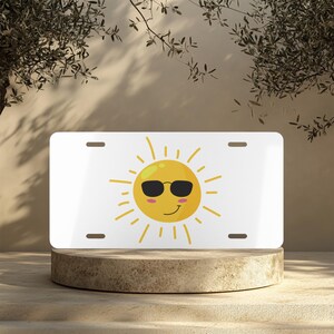Puede incluir: Placa de matrícula blanca con un sol de dibujos animados con gafas de sol. El sol es amarillo con gafas de sol negras y una cara sonriente. Los rayos amarillos se extienden desde la cara del sol. La placa tiene cuatro orificios de montaje.