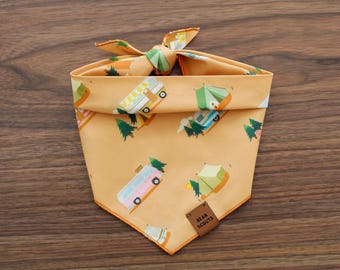 May include: A dog bandana with a camping theme. The bandana is orange with illustrations of camper vans, tents, and trees. A small brown tag reads "Bean Scouts". The bandana is tied at the top.