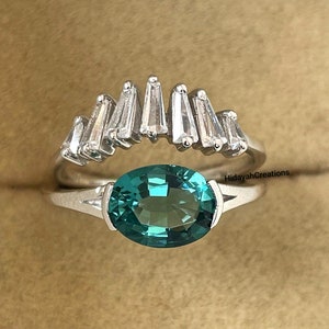 May include: A silver ring featuring a large oval teal gemstone, and a second ring with a row of baguette-cut stones. The rings are displayed against a neutral backdrop, highlighting the jewellery's design and colour.