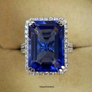 May include: A large, rectangular, sapphire-blue gemstone ring with a halo of small, clear stones. The ring's band is also adorned with smaller stones. The ring is set in a silver or white gold setting. The ring is displayed in a beige ring box.