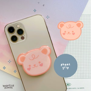 May include: A pink phone grip with a cartoon bear face design. The grip is attached to a white iPhone with a gold trim. The text "phone grip" is written on a blue circle.