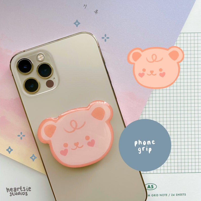 May include: A pink phone grip with a cartoon bear face design. The grip is attached to a white iPhone with a gold trim. The text "phone grip" is written on a blue circle.