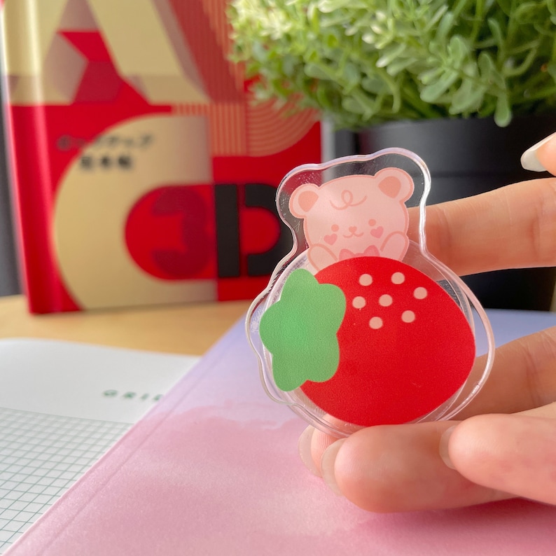 Pu&ograve; includere: Un ciondolo in plastica trasparente con un orso rosa all'interno di una fragola rossa con foglie verdi. L'orso ha orecchie rosa, un naso rosa e un papillon rosa.
