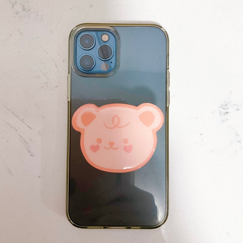 May include: A clear phone case with a dark blue back and a pink bear sticker on the back. The phone case has a camera cutout with three lenses.