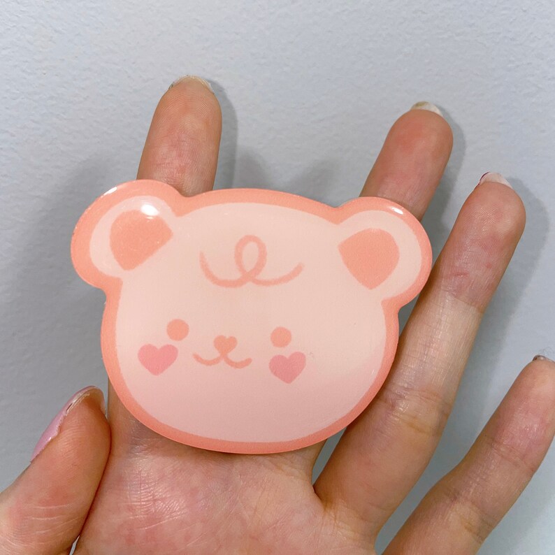 May include: A pink bear sticker with a heart on each cheek. The bear has a simple, cartoon-like design.