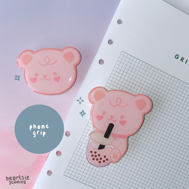 May include: Two pink bear phone grips with a cute cartoon design. One bear is holding a boba tea. The text "phone grip" is written on a blue circle.