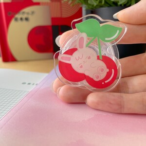 Pu&ograve; includere: Un distributore di nastro adesivo in plastica trasparente con un design a ciliegie rosse. Un coniglietto bianco con orecchie rosa e un naso rosa sta dormendo dentro la ciliegia. La ciliegia ha una foglia verde in cima.