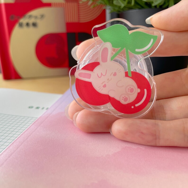 Pu&ograve; includere: Un distributore di nastro adesivo in plastica trasparente con un design a ciliegie rosse. Un coniglietto bianco con orecchie rosa e un naso rosa sta dormendo dentro la ciliegia. La ciliegia ha una foglia verde in cima.