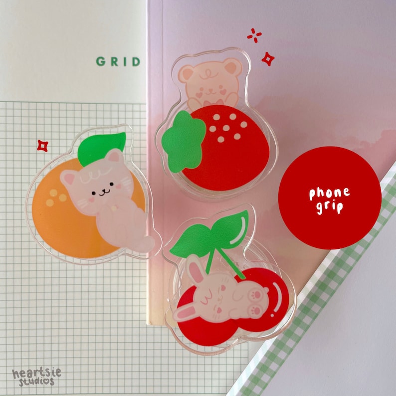 Pu&ograve; includere: Tre grip per telefono con un gatto, un coniglio e un orso. Ogni grip ha la forma di un frutto con una fragola rossa, una ciliegia rossa e un'arancia. Il testo "phone grip" &egrave; stampato su un cerchio rosso.