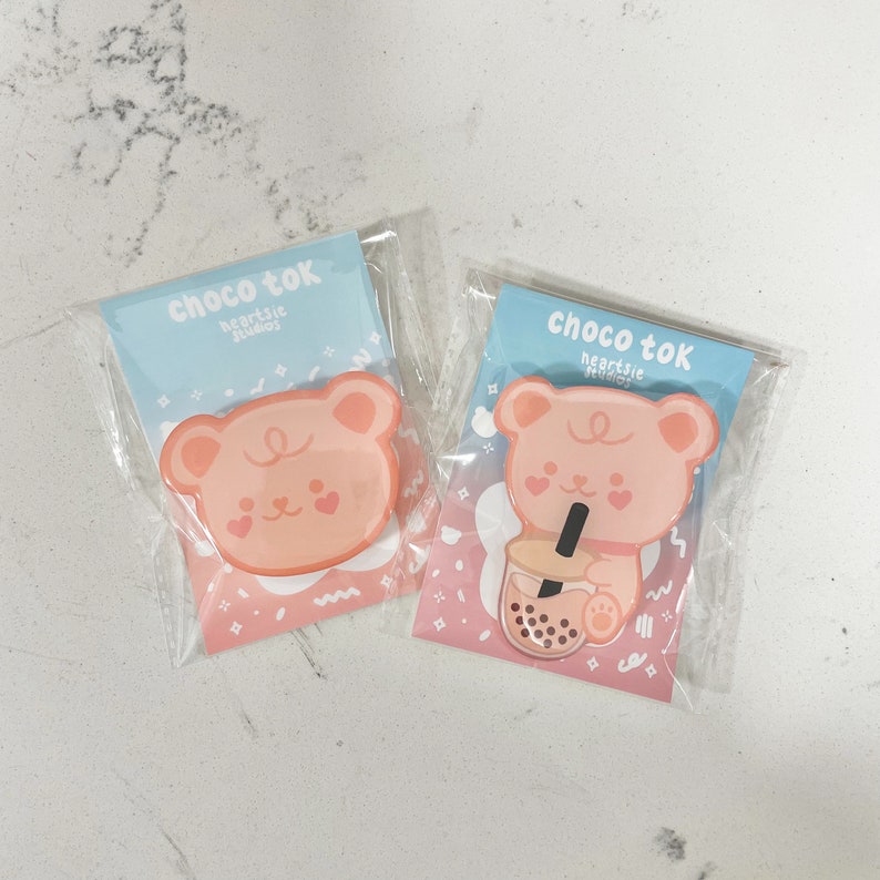 May include: Two pink and white enamel pins featuring a cartoon bear. One bear is holding a boba tea cup with tapioca pearls. The pins are packaged in clear plastic bags with pink and blue backgrounds. The text on the bags reads "choco tok heartsic studios".