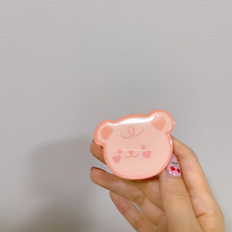 May include: A pink bear-shaped phone grip with a heart design on the cheeks. The grip is being held by a hand with cherry nail art.