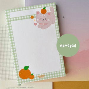May include: A green and white gingham notepad featuring a cute pink cat illustration wearing an orange on its head. The notepad has a 3mm grid and 24 sheets. The text "heartsic studios" and "A5" are printed on the notepad.