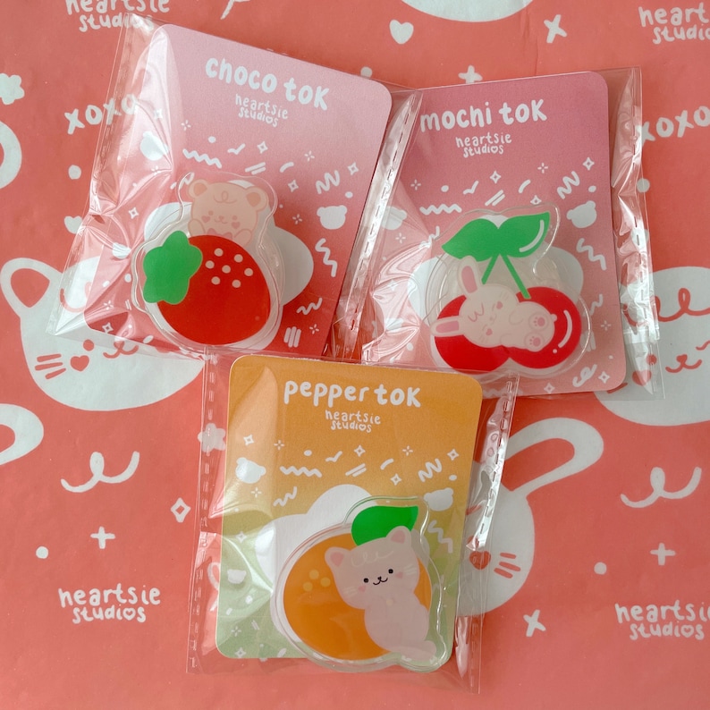 Pu&ograve; includere: Tre sacchetti di plastica trasparente, ognuno contenente un ciondolo a tema frutta con un animale dei cartoni animati. I ciondoli sono etichettati "choco tok", "mochi tok" e "pepper tok" con il testo "heartsie studios" sotto ogni etichetta.