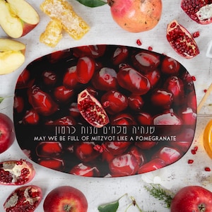 May include: A festive arrangement featuring a decorative platter with a pomegranate design, surrounded by fresh apples, pomegranate halves, honeycomb, and honey. The platter has text that reads "MAY WE BE FULL OF MITZVOT LIKE A POMEGRANATE."