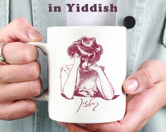 May include: White ceramic mug featuring a burgundy illustration of a woman with her head in her hands. The mug has text in English and Yiddish. The mug is held by a person.