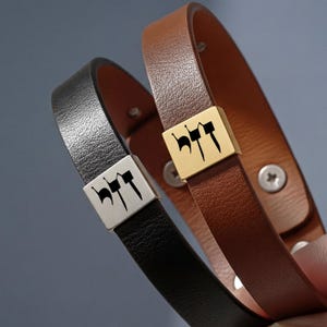 May include: Two leather bracelets, one black and one brown, are displayed. Each bracelet features a rectangular metal clasp with black lettering. The brown bracelet has a gold clasp, while the black bracelet has a silver clasp. The bracelets are fastened with a screw.