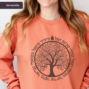 May include: A terracotta-coloured sweatshirt with a brown tree design encircled by Hebrew text. The tree and text are in a darker shade of brown. The sweatshirt has a crew neck and long sleeves.