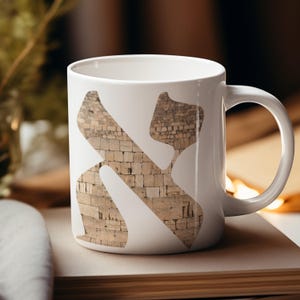 Pode incluir: Caneca de cerâmica branca com um design estilizado de uma letra hebraica preenchida com uma textura de parede de pedra. A caneca tem uma alça curva e está sobre um fundo desfocado.