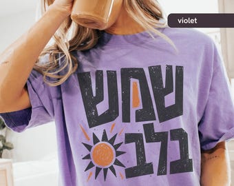 May include: A violet-colored t-shirt with a graphic design in black and orange. The design includes Hebrew text and a stylized sun. The shirt is worn by a person drinking from a clear glass.