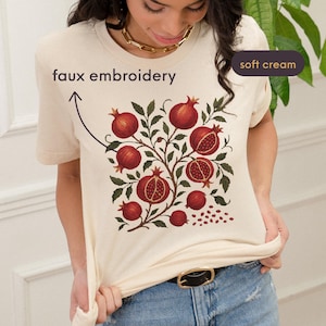 May include: A soft cream-colored t-shirt featuring a faux embroidery design of pomegranates. The design includes red pomegranates, green leaves, and the text "faux embroidery". The shirt has a round neckline and short sleeves.