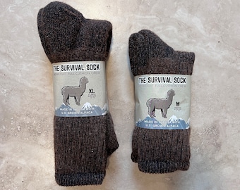 May include: Two pairs of brown alpaca wool socks. The socks are labeled "The Survival Sock" and are made in the USA with U.S. grown alpaca. The larger sock is labeled XL and the smaller sock is labeled M.