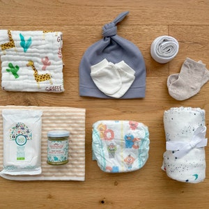 May include: A baby care package with a variety of items, including a blue and white patterned diaper, a white muslin swaddle blanket with stars, a white muslin swaddle blanket with a giraffe print, a white hat with a knot on top, a jar of baby wash, a package of baby wipes, a pair of white baby mittens, and a pair of beige baby socks.