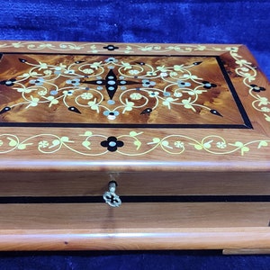 以下が含まれることがあります： 蓋に装飾的な花の象嵌が施された木製の箱。箱には鍵穴と真鍮製の鍵が付いています。箱は暗い木でできており、明るい木の縁取りがされています。