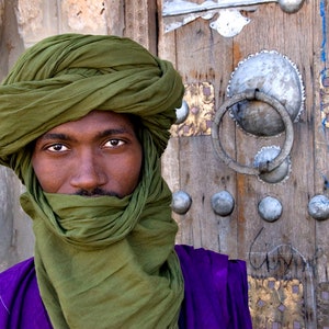 May include: A man wearing a green turban and a purple robe stands in front of a wooden door with ornate metal hardware.
