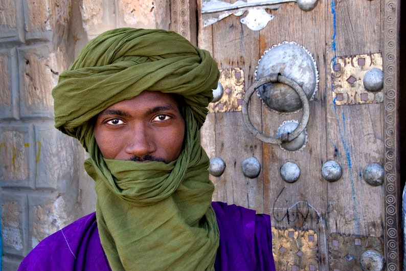 May include: A man wearing a green turban and a purple robe stands in front of a wooden door with ornate metal hardware.