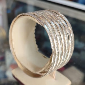 May include: A collection of silver bangles with detailed etched patterns, presented on a cream-coloured stand. The bangles are stacked, highlighting their intricate designs and reflective surface. The jewellery is displayed on a neutral background.
