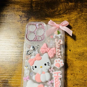 Puede incluir: Una funda de teléfono transparente con fondo de purpurina rosa. La funda presenta un diseño de Hello Kitty con dos figuras de Hello Kitty, lazos rosas y el texto "Hello Kitty" y "Sweet".