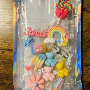 May include: A clear phone case with a rainbow, a lollipop, a gummy bear, a popsicle, a flower, and the word "Sweet" in pink letters.