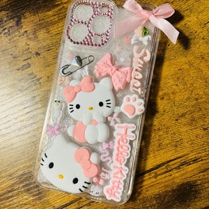 May include: A clear phone case with a pink Hello Kitty design. The case is decorated with glitter, rhinestones, bows, and a safety pin. The text "Sweet" and "Hello Kitty" is written on the case.