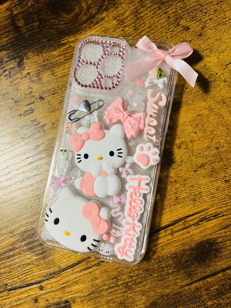 May include: A clear phone case with a pink Hello Kitty design. The case is decorated with glitter, rhinestones, bows, and a safety pin. The text "Sweet" and "Hello Kitty" is written on the case.