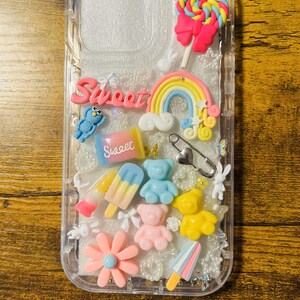 May include: A clear phone case with a glittery background and various colorful charms, including a rainbow, a lollipop, a teddy bear, a flower, and a popsicle. The word "Sweet" is written on the case in pink letters.