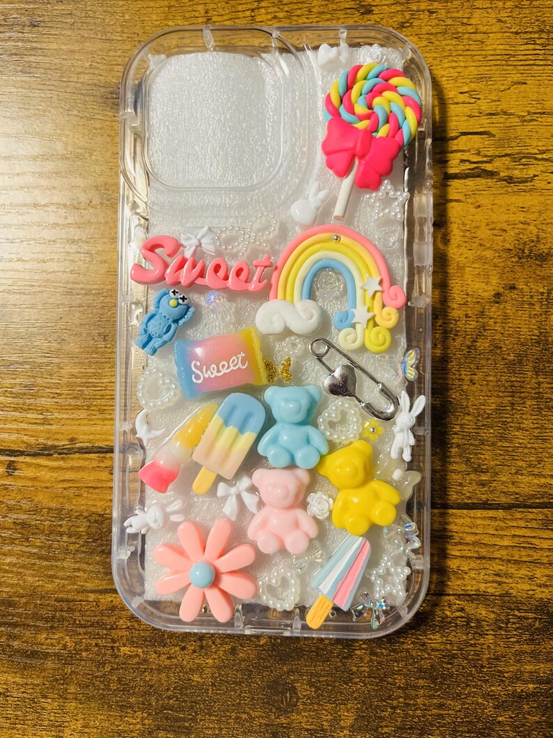 May include: A clear phone case with a glittery background and various colorful charms, including a rainbow, a lollipop, a teddy bear, a flower, and a popsicle. The word "Sweet" is written on the case in pink letters.