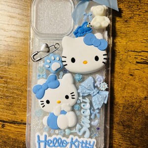 May include: A clear phone case with a glittery background and blue accents. The case features two Hello Kitty figures, a blue bow, a white teddy bear, and the text "Hello Kitty" and "Sweet".