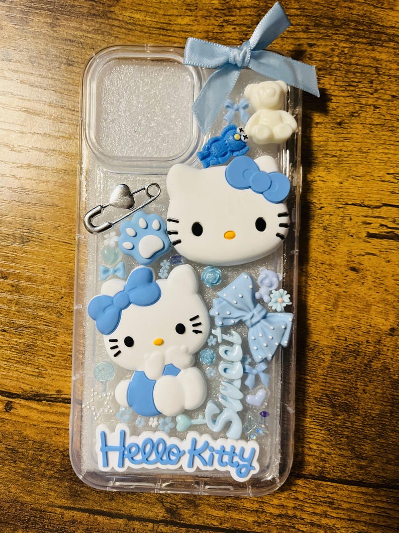 May include: A clear phone case with a glittery background and blue accents. The case features two Hello Kitty figures, a blue bow, a white teddy bear, and the text "Hello Kitty" and "Sweet".