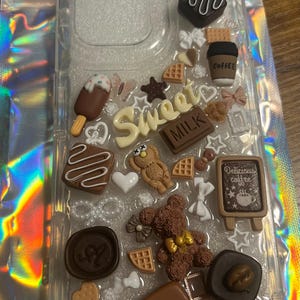 Può includere: Una custodia per telefono trasparente decorata con vari abbellimenti 3D marroni, bianchi e rosa, tra cui barrette di cioccolato, orsacchiotti, tazze da caffè e le parole "Sweet" e "Milk".