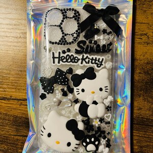 May include: A clear phone case with a black and white Hello Kitty design. The case is decorated with rhinestones, bows, and other embellishments. The text "Sweet Hello Kitty" is printed on the case.