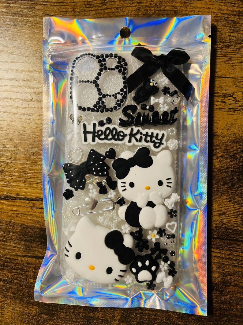 May include: A clear phone case with a black and white Hello Kitty design. The case is decorated with rhinestones, bows, and other embellishments. The text "Sweet Hello Kitty" is printed on the case.