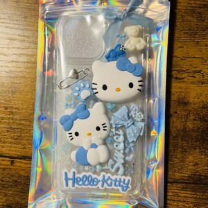 May include: A clear phone case with a blue Hello Kitty design. The case has glitter and various blue embellishments, including a bow, a teddy bear, and a paw print.