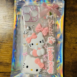 May include: A clear phone case with a pink Hello Kitty design. The case features a pink bow, a pink paw print, and a pink heart. The case also has a clear glitter background.
