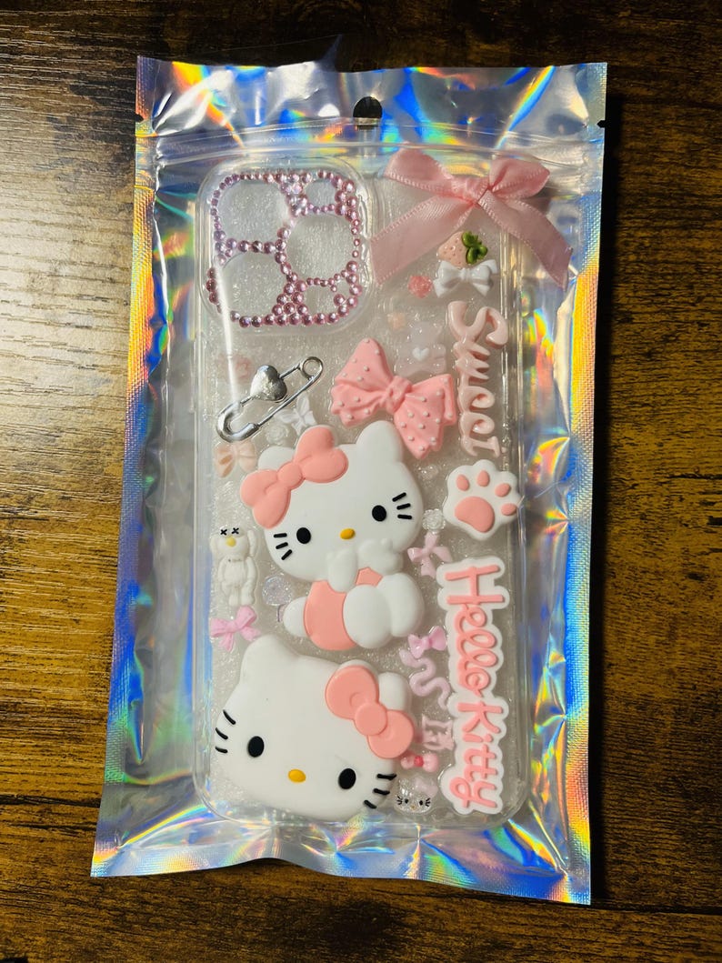 May include: A clear phone case with a pink Hello Kitty design. The case features a pink bow, a pink paw print, and a pink heart. The case also has a clear glitter background.