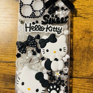 May include: A clear phone case decorated with black and white Hello Kitty characters, bows, and rhinestones. The case also features the words "Sweet" and "Hello Kitty".