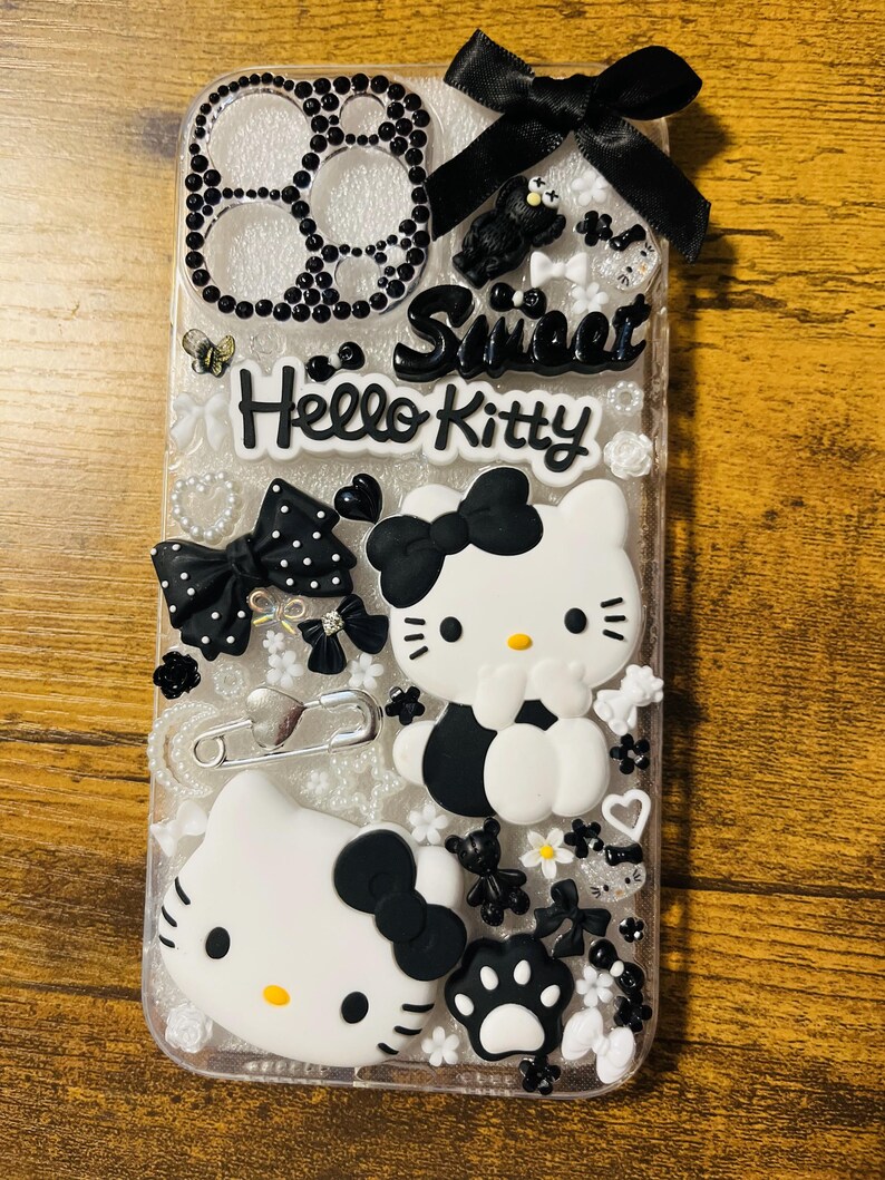 May include: A clear phone case decorated with black and white Hello Kitty characters, bows, and rhinestones. The case also features the words "Sweet" and "Hello Kitty".