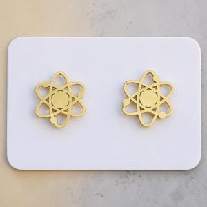 May include: A pair of gold-toned stud earrings shaped like an atom, featuring a central circular core surrounded by three intersecting elliptical orbits with small circular accents at the ends of each orbit, displayed on a white rectangular card.