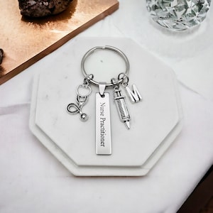 May include: Silver keychain with a silver charm that says "Nurse Practitioner" and other charms including a stethoscope, a syringe, and a letter M.